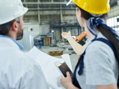 Back view portrait of female factory worker discussing  plans with mature engineer in workshop and pointing